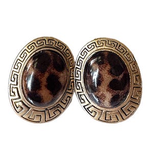May include: A pair of oval-shaped clip-on earrings. The earrings feature a gold-toned, Greek key design border surrounding a brown and black leopard print center. The earrings are set against a white background.