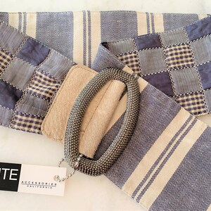 May include: A wide belt with a patchwork design in shades of blue and cream, featuring a textured oval buckle. The belt has a tag that reads "ELITE ACCESSORIES CALIFORNIA".