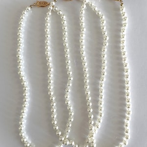 May include: Three strands of white faux pearl necklaces with gold-colored clasps. The pearls are evenly spaced and have a consistent size. The necklaces are displayed against a white background.