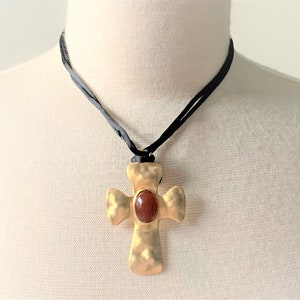 May include: A gold cross pendant with a red gemstone in the center, hanging from a black cord necklace.