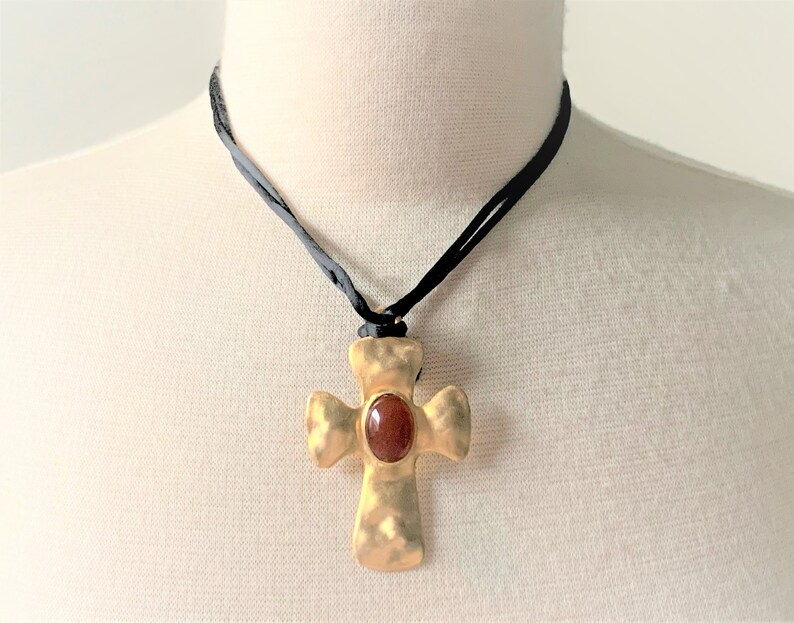 May include: A gold cross pendant with a red gemstone in the center, hanging from a black cord necklace.