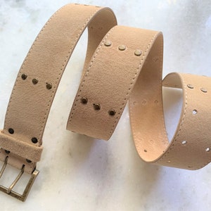 May include: A light brown suede belt with a gold metal buckle and decorative studs. The belt has a row of studs on one side and two rows of holes on the other side.