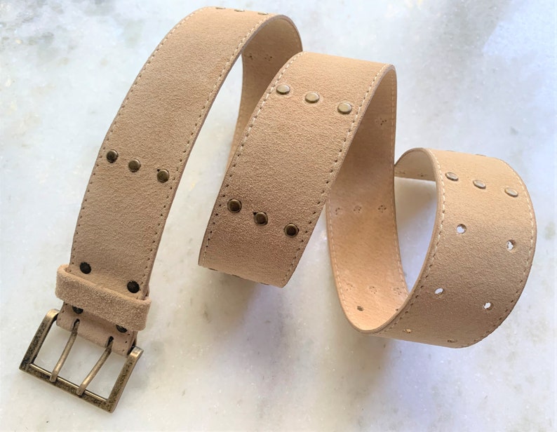 May include: A light brown suede belt with a gold metal buckle and decorative studs. The belt has a row of studs on one side and two rows of holes on the other side.