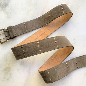 May include: A brown suede belt with gold-toned metal studs and a buckle. The belt is folded in a loop.
