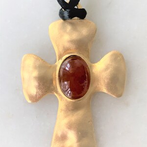 May include: A gold-toned cross pendant with a brown oval-shaped gemstone in the center. The pendant is attached to a black cord.