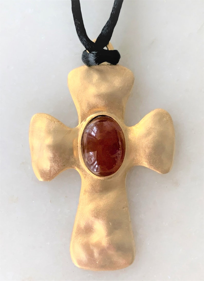 May include: A gold-toned cross pendant with a brown oval-shaped gemstone in the center. The pendant is attached to a black cord.