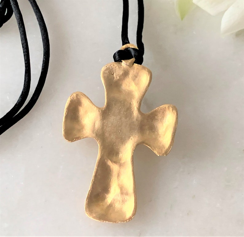 May include: A gold-toned cross pendant with a textured surface, hanging from a black cord necklace.
