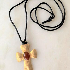 May include: A gold cross pendant with a red gemstone in the center, hanging from a black cord necklace.