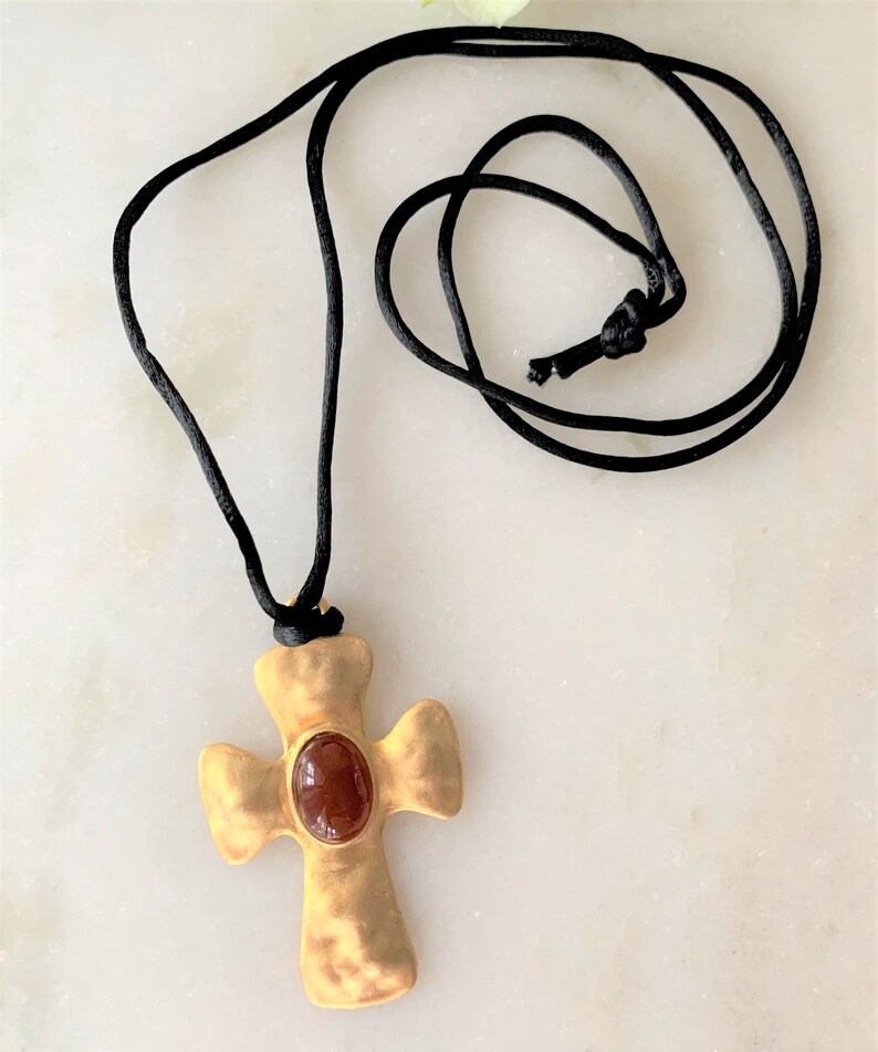 May include: A gold cross pendant with a red gemstone in the center, hanging from a black cord necklace.