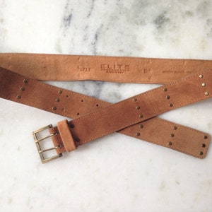 May include: A brown suede belt with gold-toned studs and a double-prong buckle. The belt is labeled "ELITE ACCESSORIES MADE IN USA" and "S/L 17".