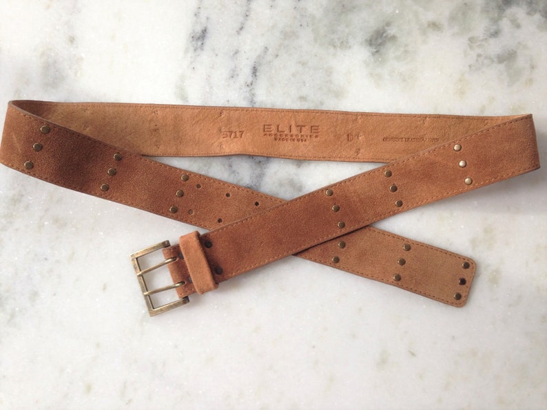 May include: A brown suede belt with gold-toned studs and a double-prong buckle. The belt is labeled "ELITE ACCESSORIES MADE IN USA" and "S/L 17".
