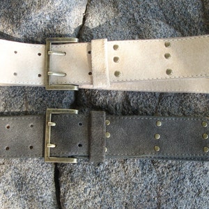 May include: Two suede belts with metal buckles and studs. The top belt is light beige and the bottom belt is dark gray.