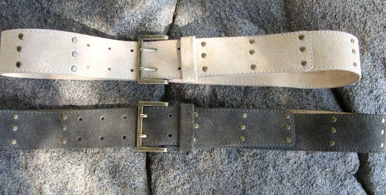 May include: Two suede belts with metal buckles and studs. The top belt is light beige and the bottom belt is dark gray.