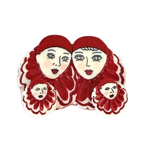 May include: A decorative brooch featuring four stylized faces with red hair and hats. The faces have pale skin, red lips, and are surrounded by a ruffled, red and white trim. The brooch has a whimsical, artistic design.