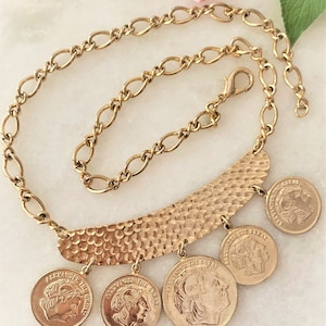 May include: Gold-toned necklace featuring a hammered bar pendant and coin charms. The necklace has a chain with oval links and a lobster clasp. Each coin charm features a profile of Alexander the Great.