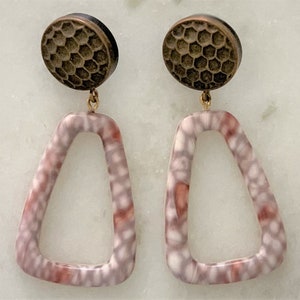 May include: A pair of earrings featuring a bronze-colored, textured top with a honeycomb pattern. The earrings have a gold-colored connecting wire and a light purple, open-frame, geometric shape with red and white accents.