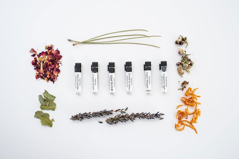 Puede incluir: Un conjunto de ocho peque&ntilde;os frascos de vidrio con tapas negras, cada uno contiene un aceite esencial diferente. Los frascos est&aacute;n dispuestos en una fila sobre una superficie blanca, con varias hierbas y flores secas esparcidas a su alrededor. Las etiquetas en los frascos dicen: "First Love", "A touch of Rose", "Mint Revival", "Kissed by Citrus", "Forest Whisper", "Tangerine Apple", "Earth Essence", y "Bar of Lavender".