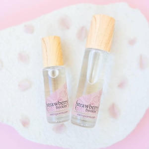 May include: Two clear glass bottles with wooden caps, labeled "strawberry freckles" with a strawberry design, rest on a white textured surface with pink petals. The bottles are filled with a clear liquid.