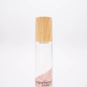 May include: A clear glass perfume bottle with a light wood cap. The label features the words "strawberry freckles" in a pink design with strawberry illustrations. The website inklingscents.com is also visible.