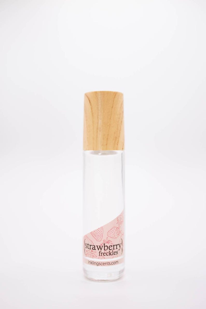 May include: A clear glass perfume bottle with a light wood cap. The label features the words "strawberry freckles" in a pink design with strawberry illustrations. The website inklingscents.com is also visible.