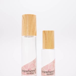 May include: Two clear glass perfume bottles with wooden caps. The bottles have a pink label with the words "strawberry freckles" and strawberry illustrations. The website inklingscents.com is printed on the label.