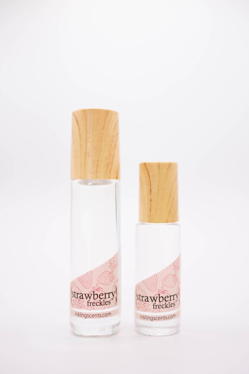May include: Two clear glass perfume bottles with wooden caps. The bottles have a pink label with the words "strawberry freckles" and strawberry illustrations. The website inklingscents.com is printed on the label.