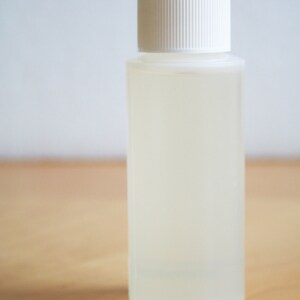 以下が含まれることがあります： 白のリブ付きキャップが付いた透明な円筒形のボトル。ボトルには、薄く半透明の液体が入っています。背景は、明るい灰色の壁と木製の表面です。
