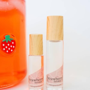 May include: Two clear glass roller bottles with wooden caps, labeled "strawberry freckles" with a strawberry design. A large glass jar with a strawberry sticker is visible in the background. The bottles are filled with a pink liquid.