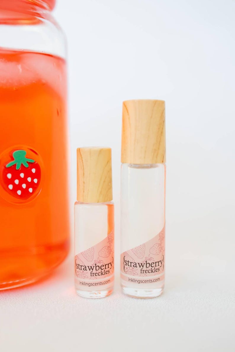 May include: Two clear glass roller bottles with wooden caps, labeled "strawberry freckles" with a strawberry design. A large glass jar with a strawberry sticker is visible in the background. The bottles are filled with a pink liquid.