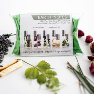 Puede incluir: Una bolsa verde que contiene un conjunto de siete peque&ntilde;os frascos de vidrio con tapas plateadas. Los frascos est&aacute;n llenos de flores y hierbas secas. La bolsa tiene una etiqueta blanca con el texto "EARTH NOTES 100% Pure Essential and Fragrance Oils Alcohol free * Chemical Free * Cruelty Free Full Roll-on 40&euro; Mini Roll-on 25&euro; inkling scents.com info@inklingscents.com". La imagen tambi&eacute;n muestra flores de lavanda secas, p&eacute;talos de rosa secos y una ramita verde.