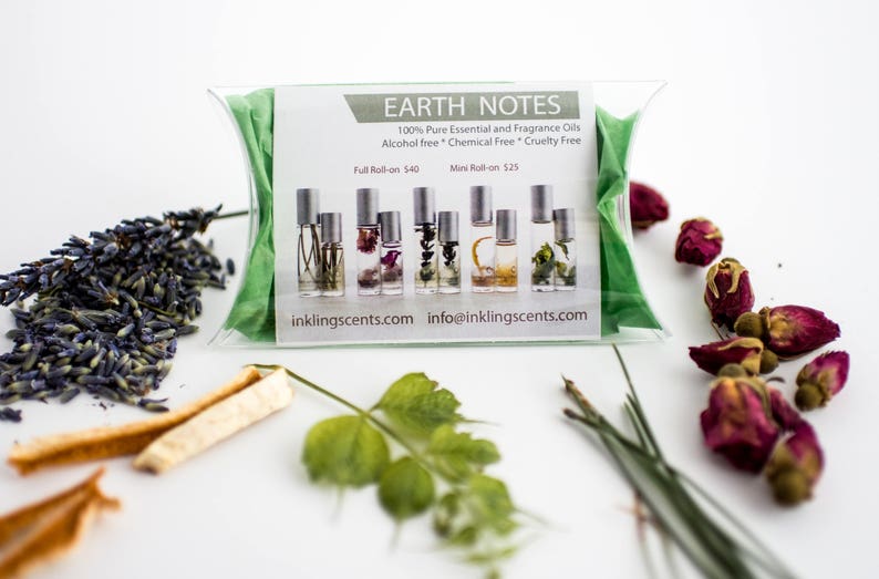 Puede incluir: Una bolsa verde que contiene un conjunto de siete peque&ntilde;os frascos de vidrio con tapas plateadas. Los frascos est&aacute;n llenos de flores y hierbas secas. La bolsa tiene una etiqueta blanca con el texto "EARTH NOTES 100% Pure Essential and Fragrance Oils Alcohol free * Chemical Free * Cruelty Free Full Roll-on 40&euro; Mini Roll-on 25&euro; inkling scents.com info@inklingscents.com". La imagen tambi&eacute;n muestra flores de lavanda secas, p&eacute;talos de rosa secos y una ramita verde.