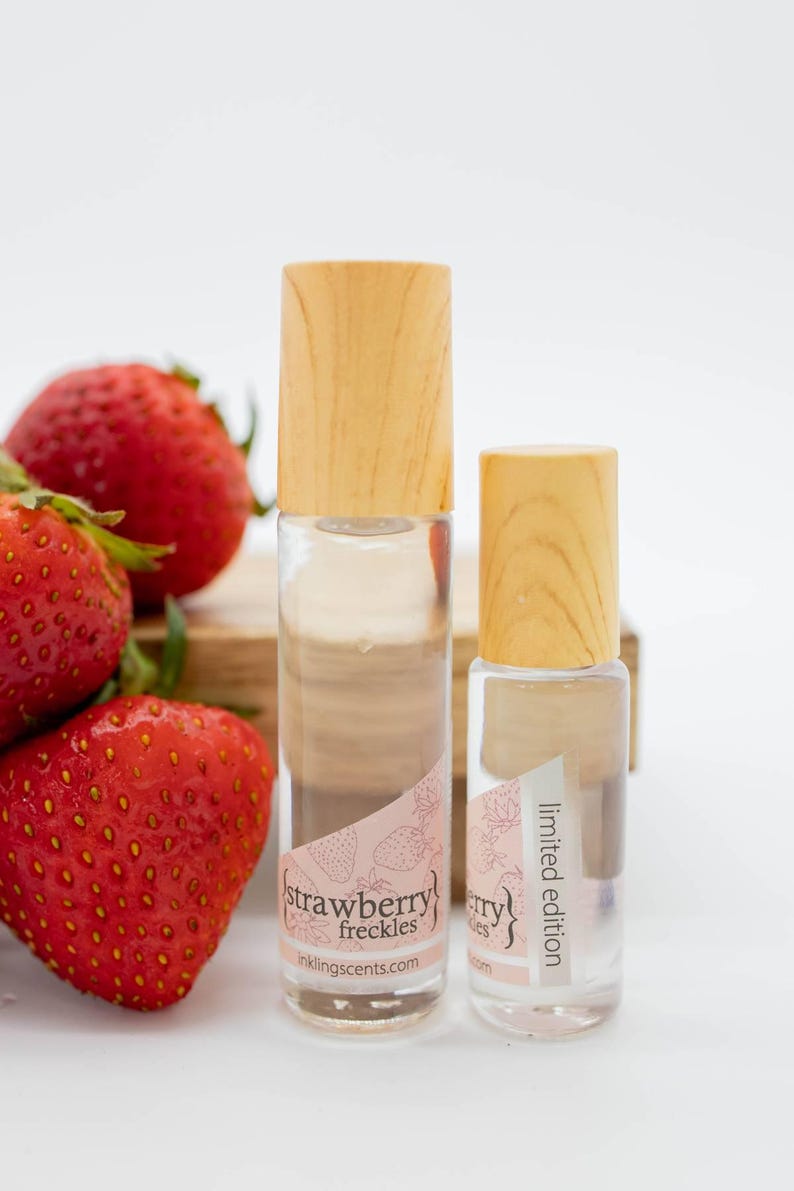 May include: Two clear glass roll-on bottles with wooden caps, labeled "strawberry freckles" and "limited edition". Fresh strawberries are in the background. The bottles have a pink label with a strawberry design.