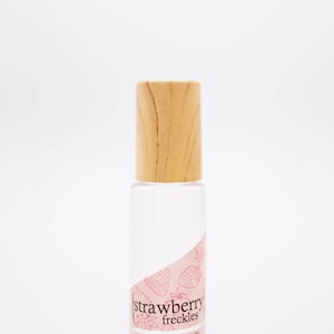 May include: A clear glass bottle with a light wood cap. The bottle has a pink label with strawberry illustrations and the text "strawberry freckles" and "inklingscents.com".