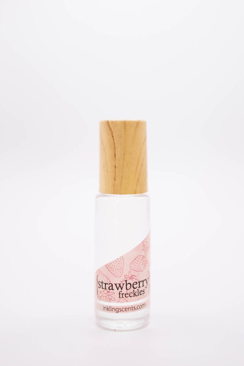May include: A clear glass bottle with a light wood cap. The bottle has a pink label with strawberry illustrations and the text "strawberry freckles" and "inklingscents.com".