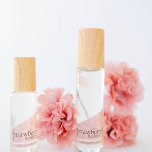 May include: Two clear glass perfume bottles with wooden caps, labeled "strawberry freckles." The bottles are next to pink flowers. The website inklingscents.com is printed on the label.