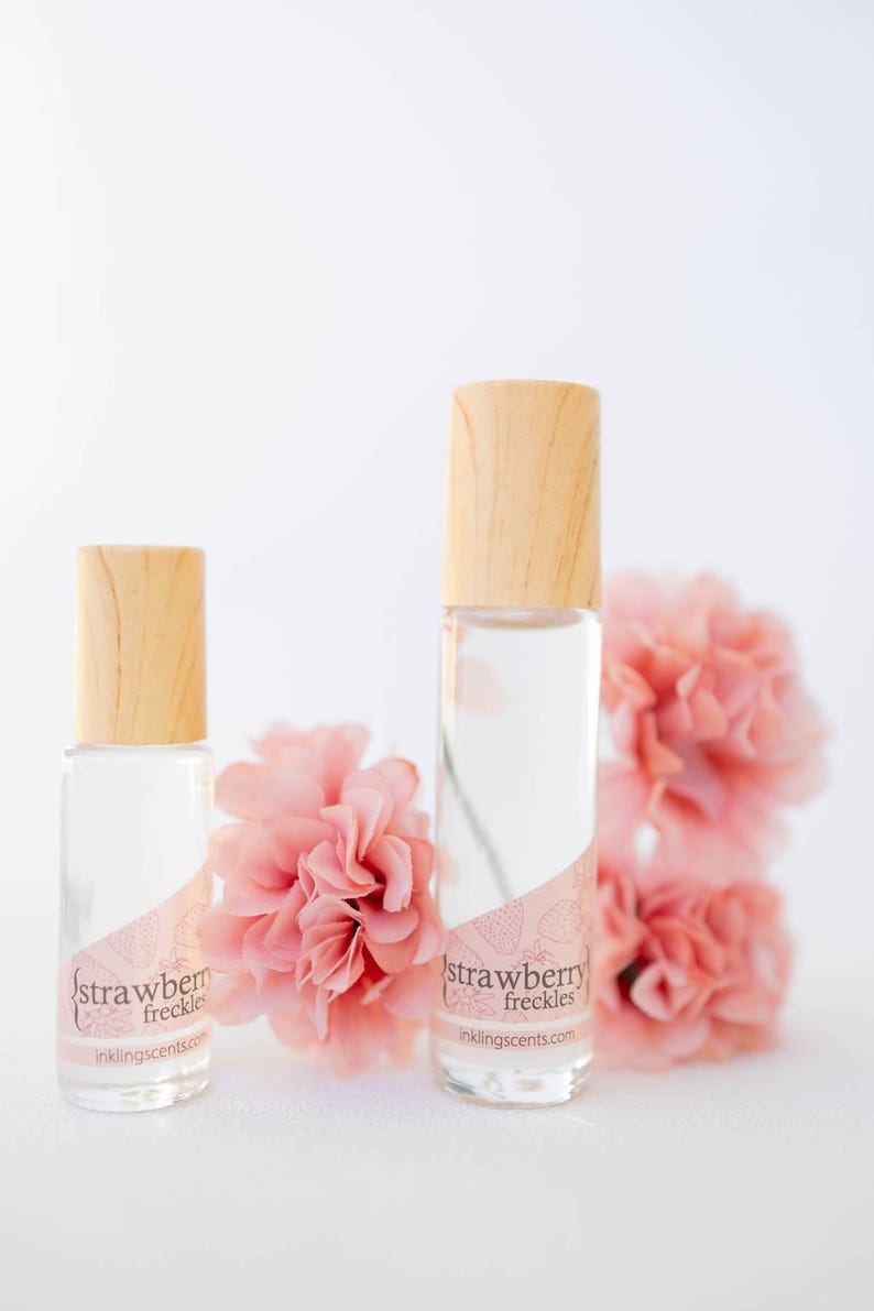May include: Two clear glass perfume bottles with wooden caps, labeled "strawberry freckles." The bottles are next to pink flowers. The website inklingscents.com is printed on the label.
