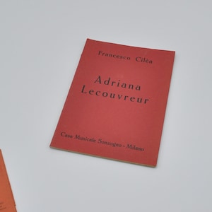 Può includere: Un libretto rosso intitolato "Adriana Lecouvreur" di Francesco Cilea. La copertina include anche il testo "Casa Musicale Sonzogno - Milano". Un altro libretto arancione è parzialmente visibile.