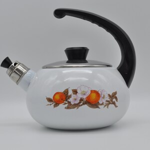 Può includere: Teiera in smalto bianco con manico e coperchio neri. La teiera ha un design floreale con frutta arancione e fiori bianchi.