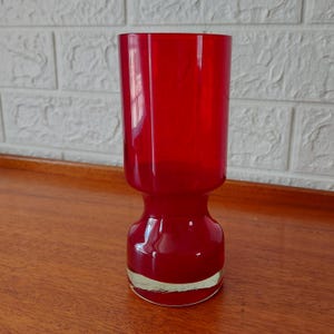May include: A tall, cylindrical red glass vase with a smooth, glossy finish. The vase features a distinct, narrow neck that flares out into a wider, rounded base, with a clear, thick glass bottom that provides a stable foundation for the vessel.