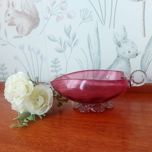 May include: A deep pink glass serving dish with a handle and a decorative base. The dish has a curved shape and sits on a base with a floral design. The handle is round and clear. The background features a floral wallpaper and a bouquet of white flowers.