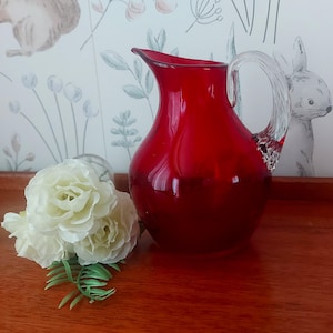 May include: A striking red glass jug with a clear glass handle. The jug has a curved spout and a rounded body. A bouquet of white flowers with green leaves sits in front of the jug. The background features a floral wallpaper.