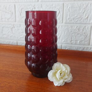 May include: A cylindrical ruby red glass vase featuring a repeating embossed circular bubble pattern across its exterior surface, accompanied by a single white artificial flower blossom placed at the base of the vessel.
