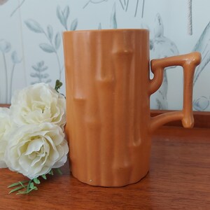 May include: Orange ceramic mug with a tree trunk design. The mug has a unique handle and a textured surface. The mug is on a wooden surface with white flowers in the foreground.