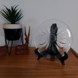 May include: A clear glass plate with a raised daisy design. The plate is on a black stand and a wooden surface.