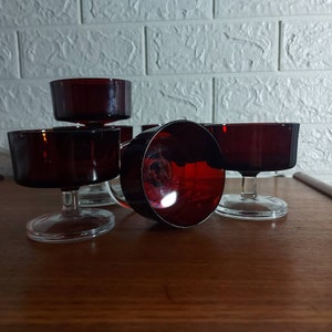 May include: A collection of six ruby red dessert glasses with stems. The glasses feature a broad, shallow bowl and a clear, slender stem. They are displayed on a dark wooden surface, with a white brick wall in the background.