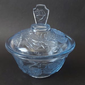 以下が含まれることがあります： 蓋付きの薄い青色のガラス製の砂糖入れ。ボウルと蓋は花柄で飾られています。蓋には盾の形をした装飾的なノブが付いています。