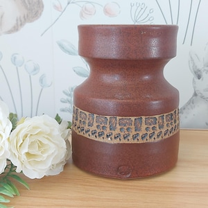May include: A brown ceramic vase with a textured surface and a decorative band of rectangular patterns. The vase has a unique hourglass shape and is suitable for home decor.