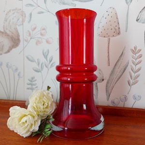 May include: A striking red glass vase with a flared top and a rounded middle section. The vase is set on a wooden surface, accompanied by two cream roses and a patterned wallpaper. The vase is approximately 30 cm tall.
