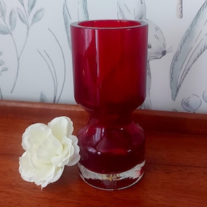 May include: A deep red glass vase with a distinctive hourglass silhouette. The vase features a broad top and a flared base. A white flower with multiple petals is positioned beside the vase. The vase rests on a wooden surface.