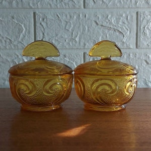 May include: Two amber glass jars with lids. The jars have a rounded shape with a swirl pattern and a small, half-moon shaped handle on the lid. The jars are on a wooden surface.